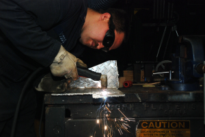 Ein Schweißer in Schutzbrille und Handschuhen bedient eine Schweißmaschine an einem Metallstück, mit einer Warntafel und verstreuten Werkzeugen im Hintergrund.