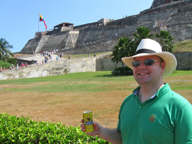 Eine Person in einem grünen T-Shirt und Brille steht links und hält eine Bierdose, mit Pflanzen im Hintergrund und einem Schloss mit Menschen und einer Fahne im Hintergrund.