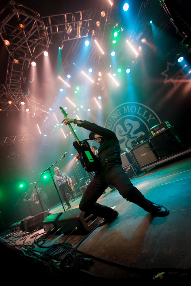 Ein musikalisches Konzert mit drei Performern auf der Bühne: zwei Gitarristen rechts mit Mikrofonen davor, ein Hintergrund mit dem Wort "MOLLEY" und zwei Schlangenlogos sowie Scheinwerfer darüber.