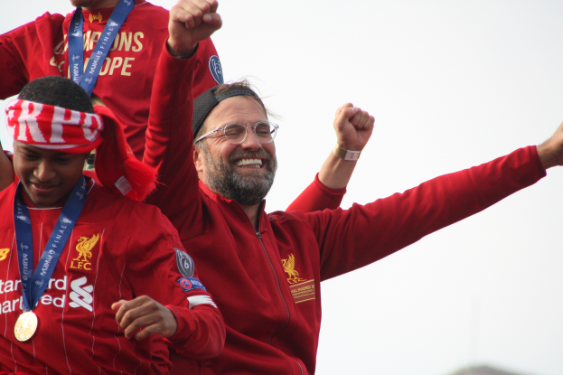 Liverpool-Trainer Jürgen Klopp feiert mit seinen Spielern den Sieg in der UEFA Champions League, während er eine rote Jacke trägt und von zwei anderen Personen mit Medaillen umgeben ist. Der Himmel ist im Hintergrund zu sehen.