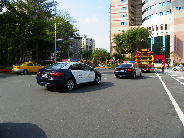 Ein Polizeiwagen fährt auf einer Straße mit hohen Gebäuden, Bäumen, Laternen, Ampeln und Menschen unter einem bewölktem Himmel.