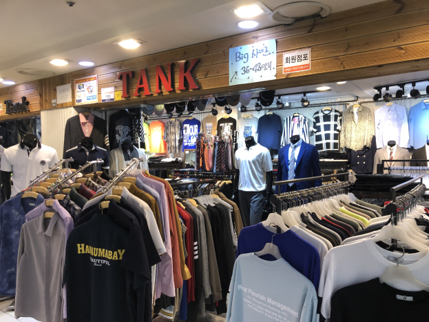 Ein modernes Bekleidungsgeschäft mit zahlreichen Hemden auf der Auslage, Schaufensterpuppen, die aktuelle Outfits präsentieren, beleuchtete Texttafeln an den Wänden und Deckenleuchten.