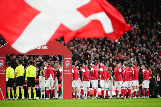 Gruppe von Menschen auf einem Fussballfeld mit einer roten und weissen Flagge im Vordergrund, einem Bogen mit der Aufschrift "Bayern München vs Bayern München Betting Tips & Preview" im Hintergrund und einer grossen Menge im Stadion.
