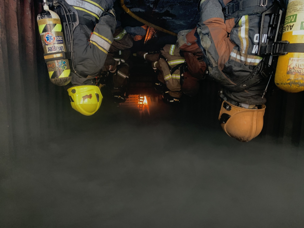 Feuerwehrleute mit Helmen und Jacken mit Atemschutz arbeiten in einem raucherfüllten Tunnel, mit einer Lichtquelle im Hintergrund.