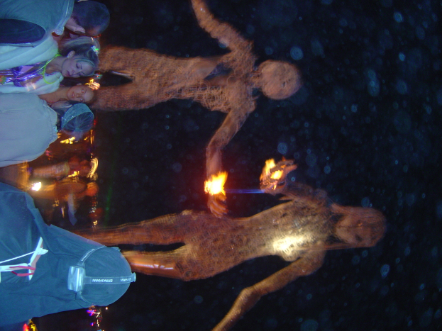 Eine Gruppe von Menschen auf der linken Seite des Bildes, mit brennenden Statuen dahinter und Lichtern im Hintergrund.