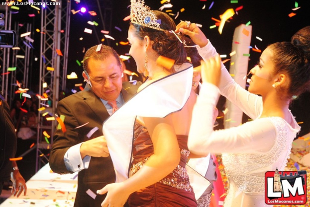 Ein Mädchen auf einer Bühne mit einer Krone, mit einem anderen Mädchen, das sie von hinten anpasst, und einem Mann in einem schwarzen Anzug, der vor ihr die Hand schüttelt, vor einem dunklen Hintergrund mit Metallrahmen.