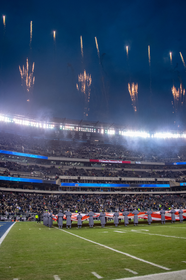 Ein Fußballstadion voller stehender Zuschauer unter hellen Lichtern, umgeben von textbedeckten Tafeln, mit Feuerwerk am Himmel.
