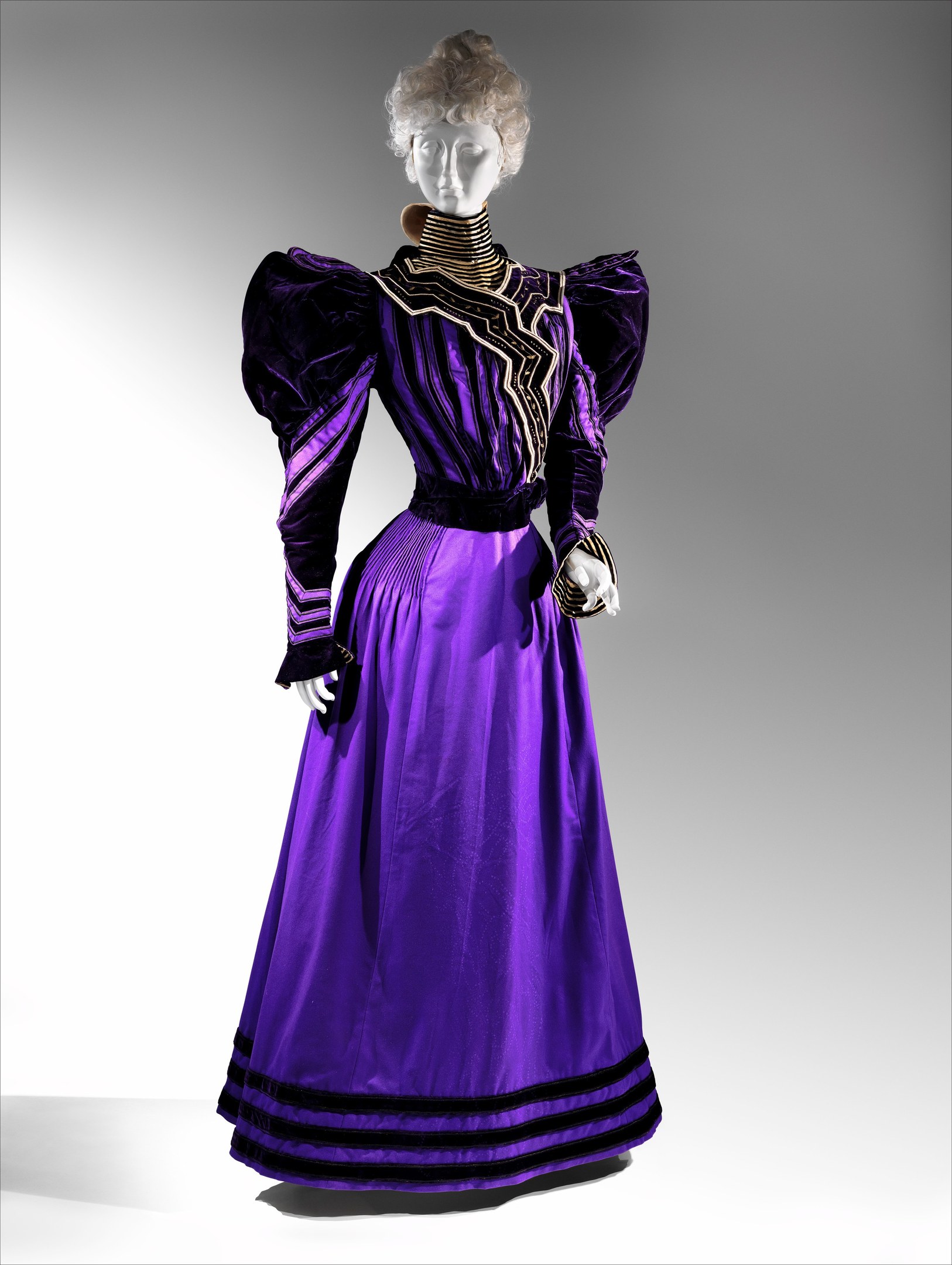 Eine Schaufensterpuppe in einem lila Abendkleid mit schwarzem und goldenem Muster, das eine eng anliegende Taille, einen langen Bodenrock mit goldener Stickerei und Perlen sowie eine Spitzenborte an der Taille hat und mit einem eleganten Dutt und zarten Ohrringen kombiniert ist.