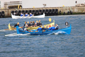 Eine Gruppe von Menschen in einem blauen und gelben Boot auf dem Wasser, die Paddel halten, mit einer Wand aus Reifen und einem Gebäude im Hintergrund, was eine Regatta vermuten lässt.