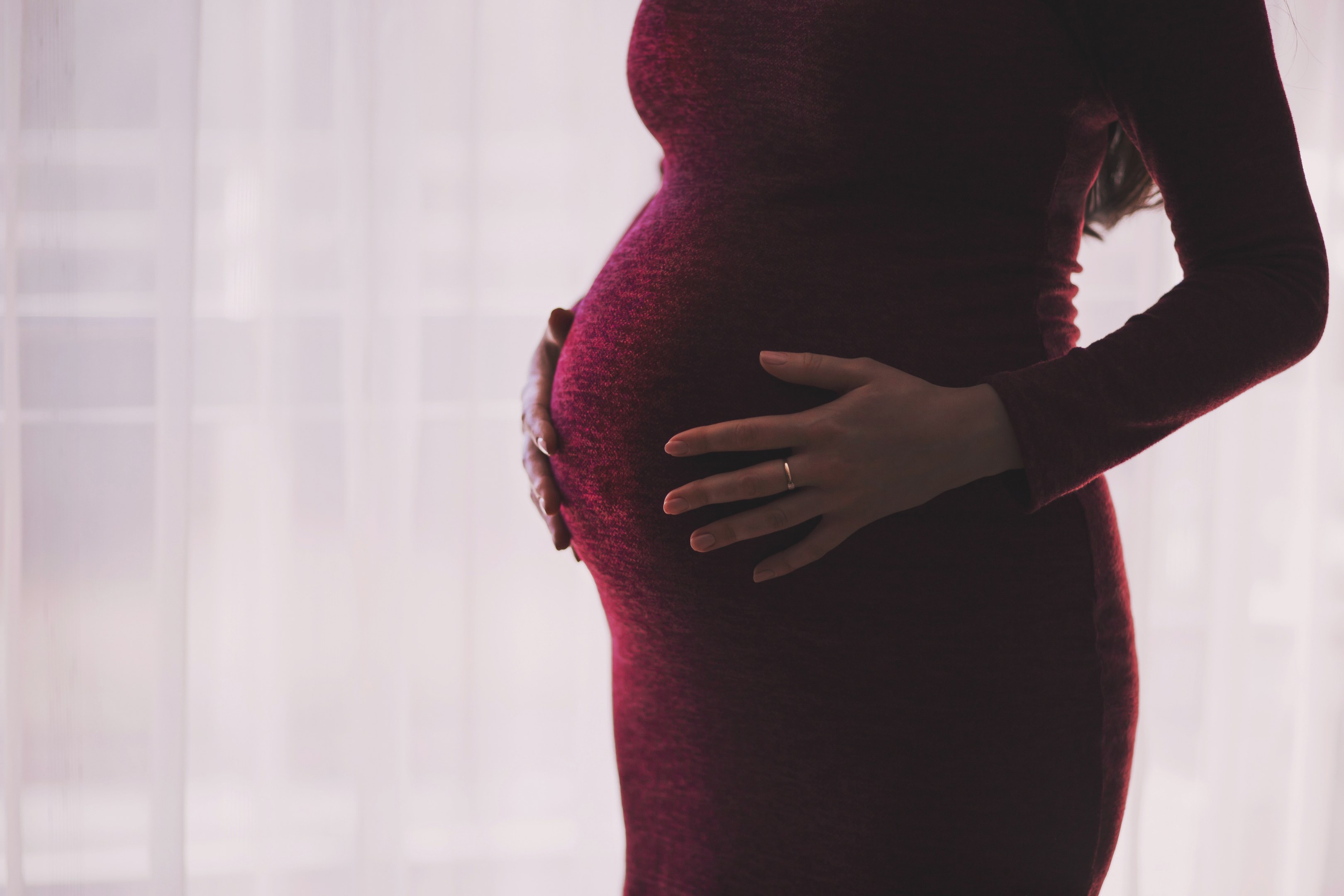 Eine schwangere Frau in einem roten Kleid steht neben einem Fenster, Hände auf dem Bauch, mit einem sanft unscharfen Hintergrund.