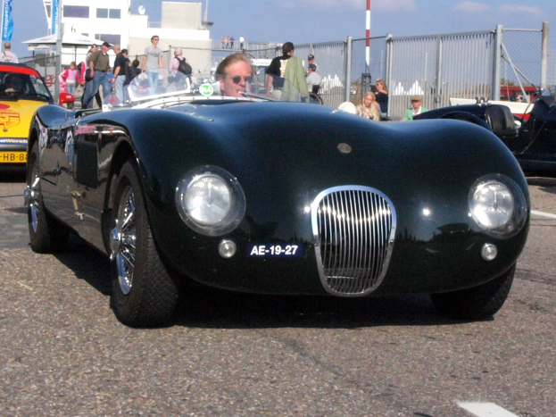 Ein schwarzer Sportwagen auf einem Parkplatz mit einer Person auf dem Fahrersitz, Menschen in der Nähe, einem Zaun, Pfosten, einem Gebäude und einem bewölkten Himmel im Hintergrund.