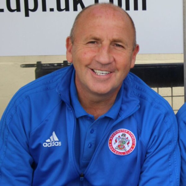 Zwei strahlende Männer in einem Raum, einer in einem blauen Jackett, der wahrscheinlich als Trainer des Accrington Stanley Football Clubs identifiziert wurde, mit einer Tafel an der Wand dahinter.