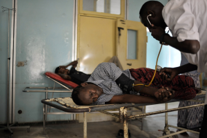 Ein Mann liegt auf einer Krankenhausliege, während ein Arzt seinen Blutdruck mit einem Stethoskop überprüft; zwei weitere liegen auf Tragen im Hintergrund bei Türen und einer Wand.