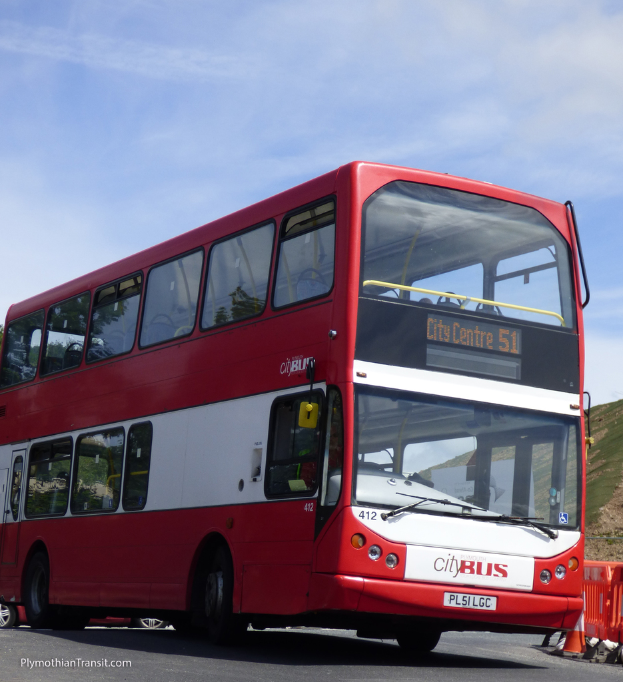 Ein roter Doppeldecker-Stadtbus ist auf der Stra√üe unterwegs mit einem Verkehrskegel davor und einem Hügel im Hintergrund.
