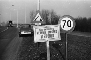 Ein Schwarz-Weiß-Foto eines Geschwindigkeitslimitschildes am Straßenrand mit einem vorbeifahrenden Auto, Bäumen, Pfählen und einem klaren Himmel im Hintergrund.