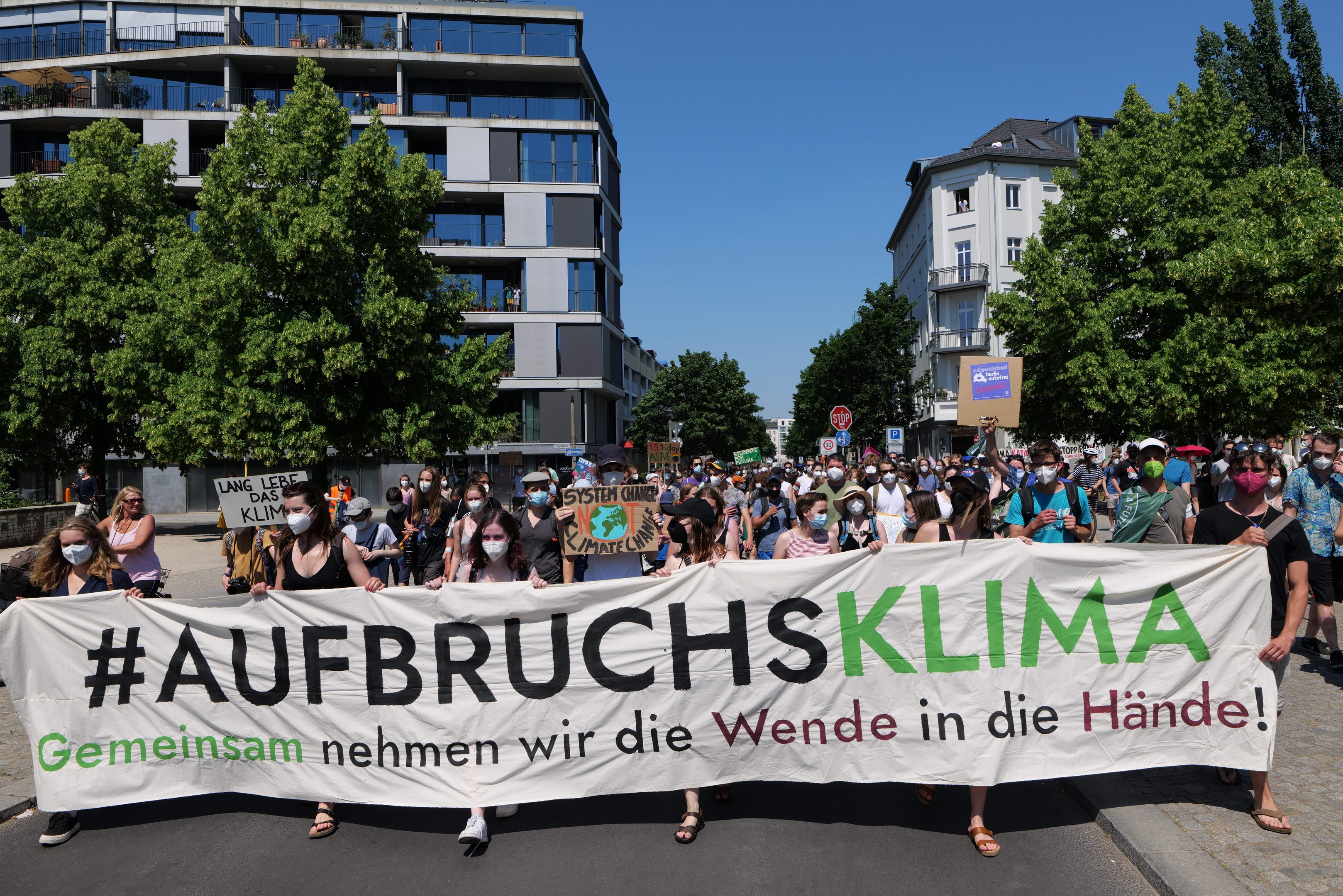 Eine Gruppe von Menschen mit Masken, die ein Banner mit der Aufschrift 'Aufbruchsklima' vor einem Gebäude mit Bäumen und einem klaren Himmel halten.
