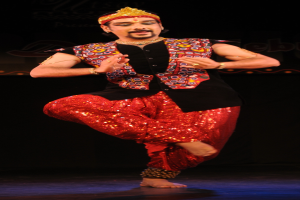 Ein Mann in roter und schwarzer Kleidung tanzt auf einer Bühne mit dunklem Hintergrund, sein Gesicht ist sichtbar mit geschlossenen Augen und leicht geöffnetem Mund.