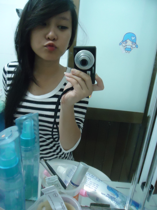 Eine Frau steht vor einem Schminktisch mit Kosmetikartikeln und macht ein Foto mit einer Kamera, wobei eine Tür im Hintergrund zu sehen ist.