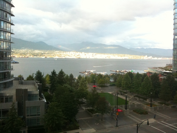Das Bild zeigt eine Küstenlandschaft mit Gebäuden und Bäumen auf beiden Seiten, einem Weg und Pflanzen unten, Booten in der Mitte, einem Strand und Bergen im Hintergrund und dem Himmel oben.