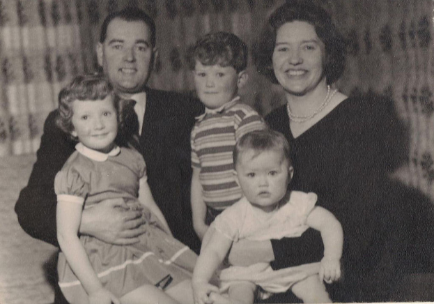 Eine Familie mit fünf Personen, mit einem Mann in Anzug und Krawatte links, einer Frau rechts und drei strahlenden Kindern in der Mitte.