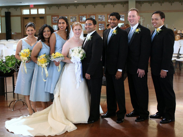 Eine Frau in einem weißen Kleid mit einem Strauß steht in der Mitte, umgeben von Frauen in blauen Kleidern und Männern in schwarzen Anzügen, die alle lächeln und drinnen mit verschiedenen Gegenständen im Hintergrund stehen.
