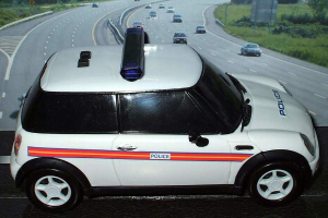 Ein weißer Polizeiwagen steht im Vordergrund mit mehreren Autos, die hinter ihm auf der Straße fahren, umgeben von Gras und Bäumen auf beiden Seiten in einer ländlichen Umgebung.