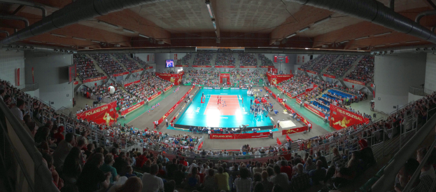 Eine große Halle mit Zuschauern bei einem Volleyballspiel, mit Bannern, Bildschirmen und Deckenbeleuchtung, sowie Geländern unten.
