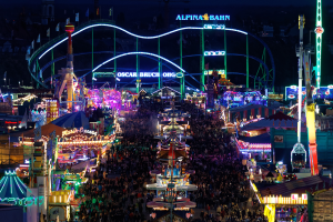 Eine große Menschenmenge steht um einen hell erleuchteten Karneval in der Nacht herum, umgeben von Vergnügungsgeräten, Pfosten, Flaggen und Namensschildern, mit einem dunklen Hintergrund, der eine festliche Atmosphäre schafft.