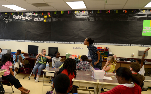 Ein Klassenzimmer mit Kindern an Tischen mit verschiedenen Gegenständen, einer Lehrerin oder einem Lehrer in der Mitte und einer Tafel im Hintergrund.