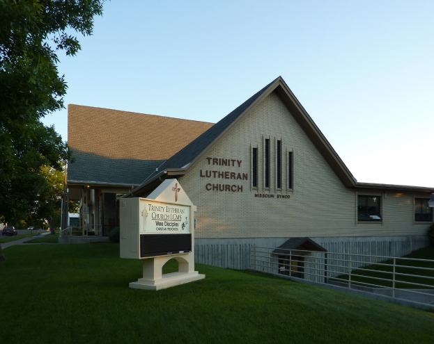 Trinity Lutheran Church in St. Louis, Missouri, ein Gebäude mit Fenstern, einem Schild, einem Metallzaun, Gras, einem Straßenpfahl, Bäumen, Fahrzeugen auf der Straße und einem bewölkten Himmel.