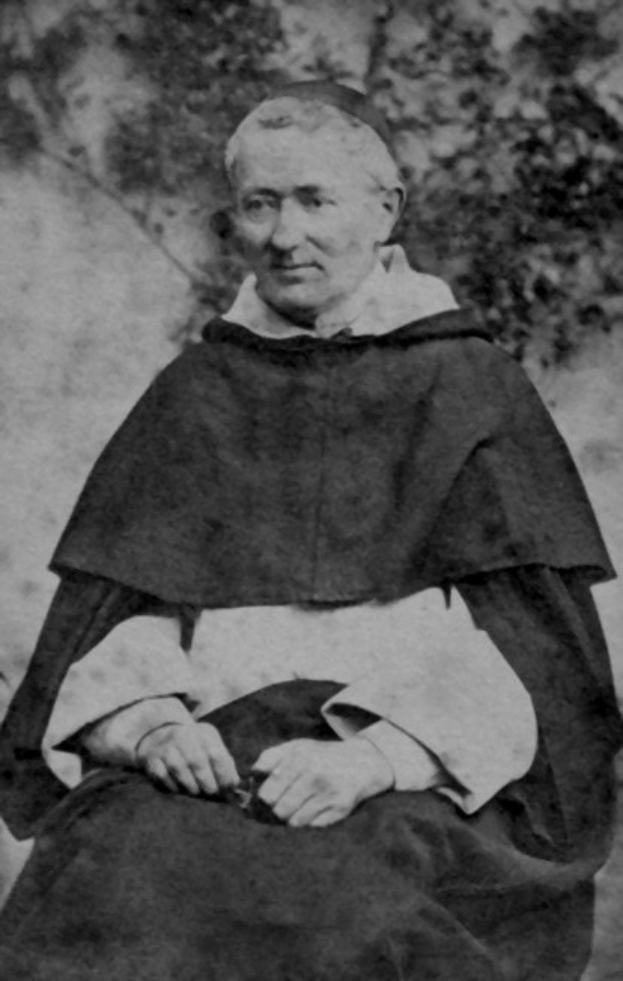 Altes Schwarz-Weiß-Foto eines Mannes in Priesterrobe vor Bäumen sitzend, in schwarzer Robe mit weißem Kragen und Manschetten, Hände zum Gebet gefaltet, von warmem Licht erhellt mit friedlichem Ausdruck.