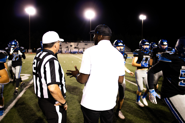 Ein Fußballtrainer in einer Mütze spricht zu seinem Team von Helmträgern auf einem Feld bei Nacht, mit Leuchtmasten und einem dunklen Himmel im Hintergrund.