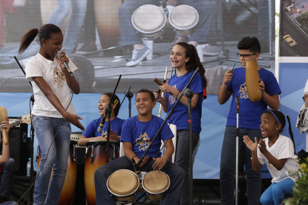 Eine Gruppe von Kindern, die auf einer Bühne Musikinstrumente spielen, mit einer Frau in der Mitte, die ein Mikrofon hält, umgeben von Mikrofonständern, einem Banner im Hintergrund und Pflanzen auf der rechten Seite.