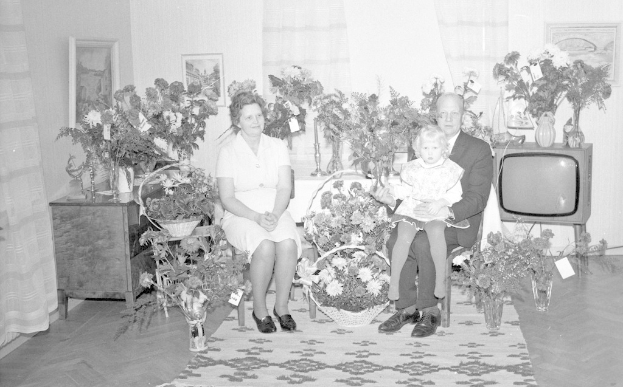 Ein Schwarz-Weiß-Foto einer Familie aus drei Personen, die auf einem Sofa vor einem Fernseher sitzen, mit Blumenvasen auf nahen Tischen und gerahmten Bildern an der Wand.