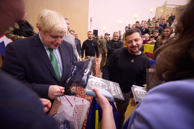 Mann in Anzug und Krawatte signiert Autogramme für eine Menschenmenge, einige halten Taschen und Bücher, mit beleuchteter Wand im Hintergrund.