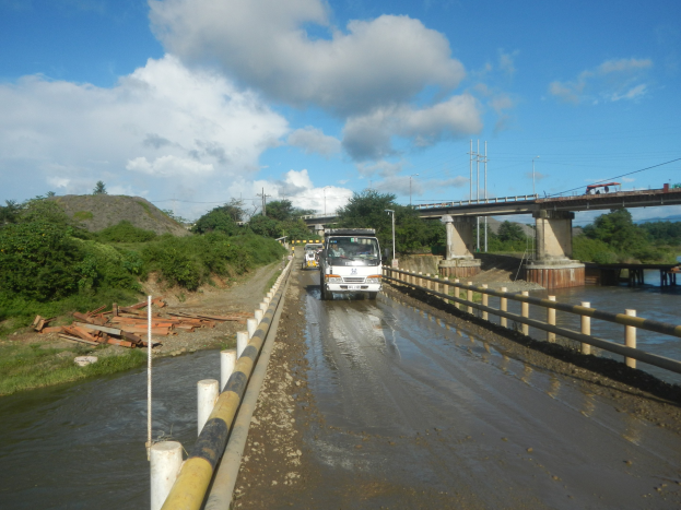 Ein Lastwagen fährt auf einer überfluteten Straße neben einer Brücke mit Geländern, mit Bäumen, Strommasten und einem bewölkten Himmel im Hintergrund.