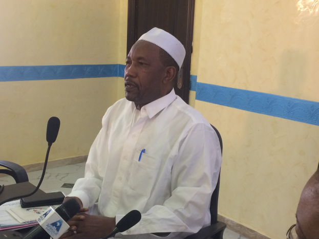 Ein Mann in einem weißen Kleid und einer Mütze sitzt an einem Tisch mit Mikrofonen und spricht, während Papiere und Gegenstände auf dem Tisch zu sehen sind, mit einer anderen Person mit Brille rechts von ihm, in einem Raum mit einer Wand und einer Tür dahinter.