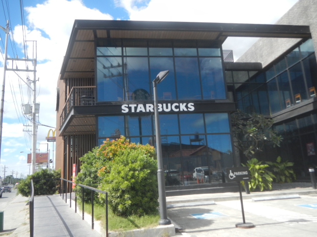 Ein Starbucks-Café an der Ecke einer Straße mit umliegenden Gebäuden, Straßeninfrastruktur, Fahrzeugen, Grünflächen und einem bewölkten Himmel.