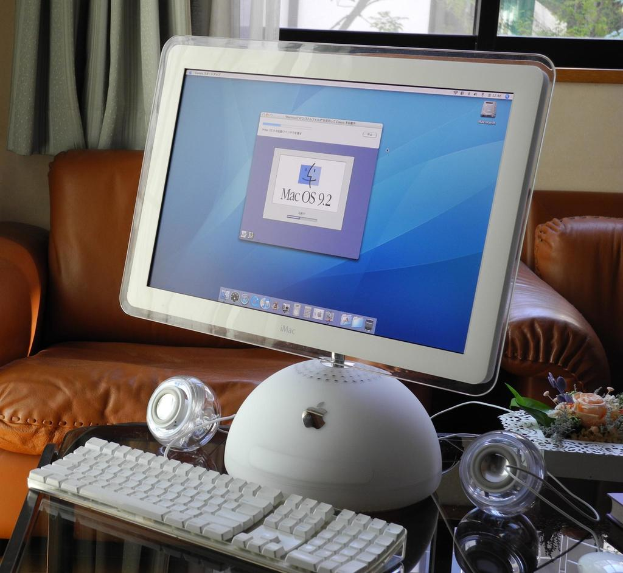 Ein Apple-Monitor, eine Tastatur und zwei Lautsprecher auf einem Schreibtisch mit zwei braunen Sofas dahinter, einem Vorhang und einem Fenster im Hintergrund.