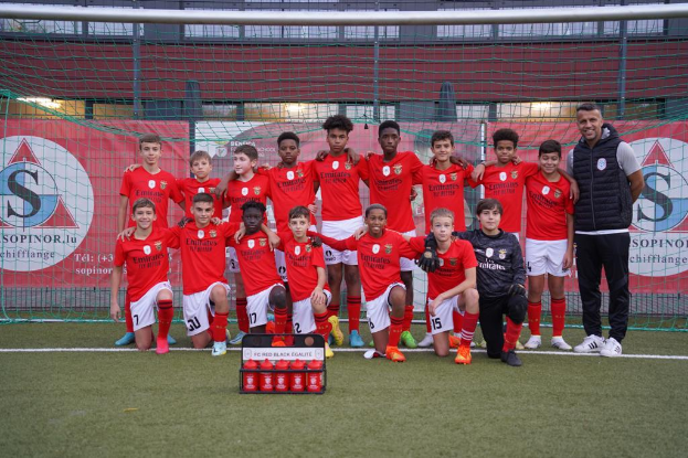 Eine Gruppe junger Männer in roten T-Shirts und schwarzen Shorts steht auf einem Fußballfeld neben einer Kiste roter Gegenstände, im Hintergrund Schilder mit der Aufschrift "FC Red Black Eglaute - Champions League".