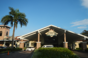 Einfahrt zu einem Resort mit Palmen vorne, umgeben von Gebäuden, Fahrzeugen auf der Straße, Verkehrskegeln, Büschen, Bäumen und einem bewölkten Himmel im Hintergrund.