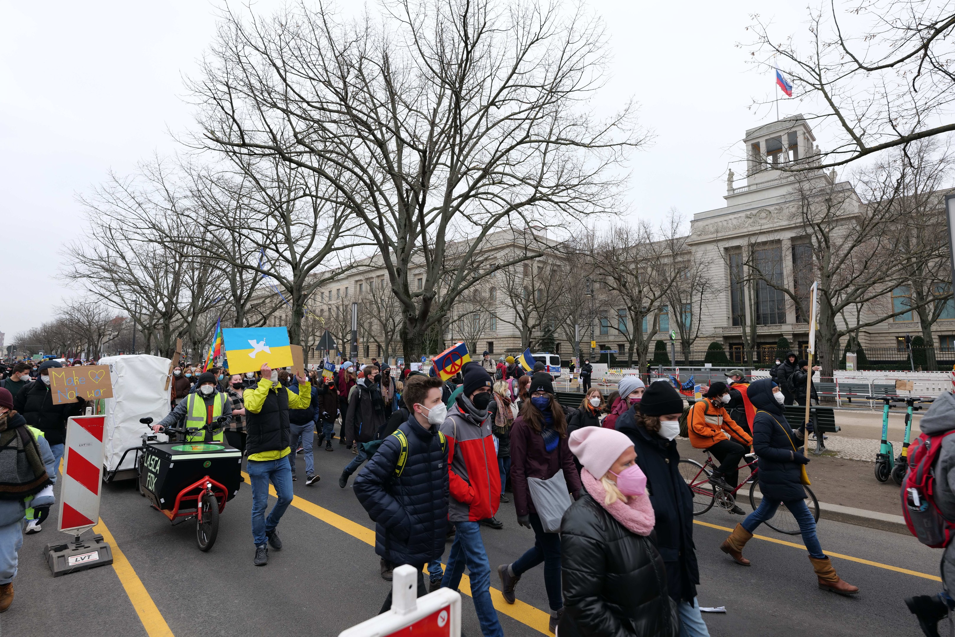 Eine große Gruppe von Menschen nimmt an einer Protestmarsch teil und hölt Schilder und Banner in den Hönden, während einige Fahrráder fahren, unter einem klaren blauen Himmel.