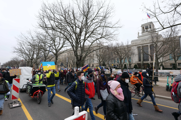 Eine große Gruppe von Menschen nimmt an einer Protestmarsch teil und hölt Schilder und Banner in den Hönden, während einige Fahrráder fahren, unter einem klaren blauen Himmel.