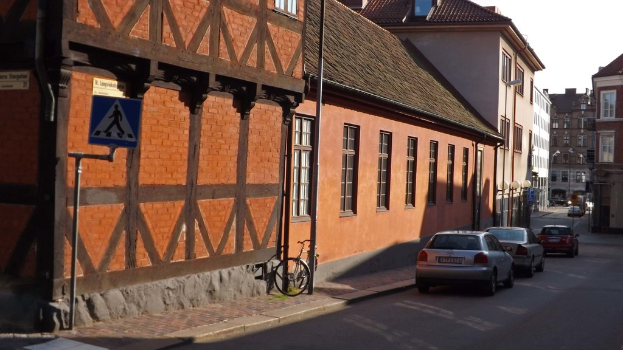 Fahrzeuge auf der Straße mit Gebäuden, Laternenmasten, Schildermasten, Fenstern und Dächern im Hintergrund und ein Fahrrad auf dem Gehweg in der Nähe eines Mastes.