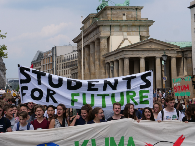 Gruppe von Schülern marschiert in Berlin mit einer bunt bemalten "Students for Future"-Schultertasche vor einer Kulisse aus Gebäuden, Bäumen und Himmel.