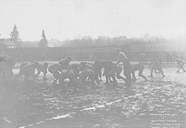 Ein Schwarz-Weiß-Foto einer Gruppe von Menschen, die auf einem Feld Fußball spielen, mit Pferden im Vordergrund und Bäumen, Gebäuden und Himmel im Hintergrund. Unten auf dem Bild steht der Text "1918-1918 Fußball an der Staatsuniversität."
