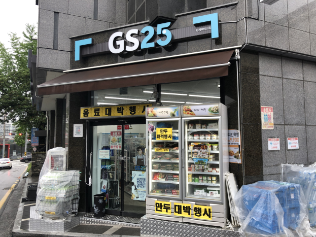 Außenansicht eines GS25-Ladens mit einem sichtbaren Schild, parkenden Autos auf der Straße, Bäumen und einem klaren blauen Himmel; drinnen, Kühlgeräte mit Lebensmitteln gefüllt.