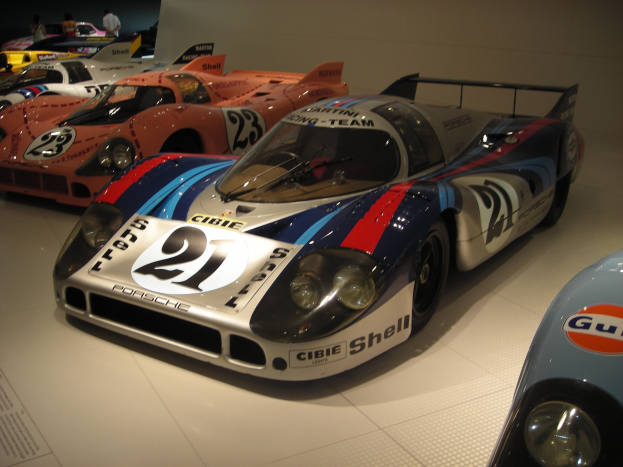Ein Porsche 917K Rennwagen auf einer Museumsausstellung umgeben von anderen Sportwagen, mit Menschen in der Nähe und einer Wand im Hintergrund; ein Schild mit Text ist links sichtbar.