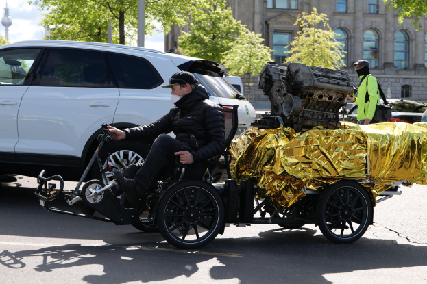 Ein Mann im Rollstuhl mit einem großen Motor auf dem Rücken, umgeben von Fahrzeugen auf einer Straße, mit Bäumen, Gebäuden, Polen und einem klaren blauen Himmel im Hintergrund.