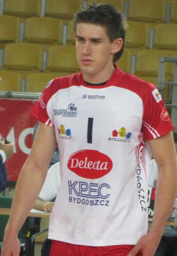 Ein Mann in rot-weißer Handballuniform steht auf einem Feld, umgeben von Zuschauern, mit einem roten Banner "Krasnoyarsk vs Bydgoszcz" im Hintergrund, entschlossen aussehend.